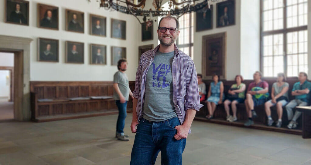 Matthew Rees steht lächelnd in dem historischen Saal des Rathauses mit Porträts an den Wänden; im Hintergrund sitzen weitere Personen auf einer Holzbank.