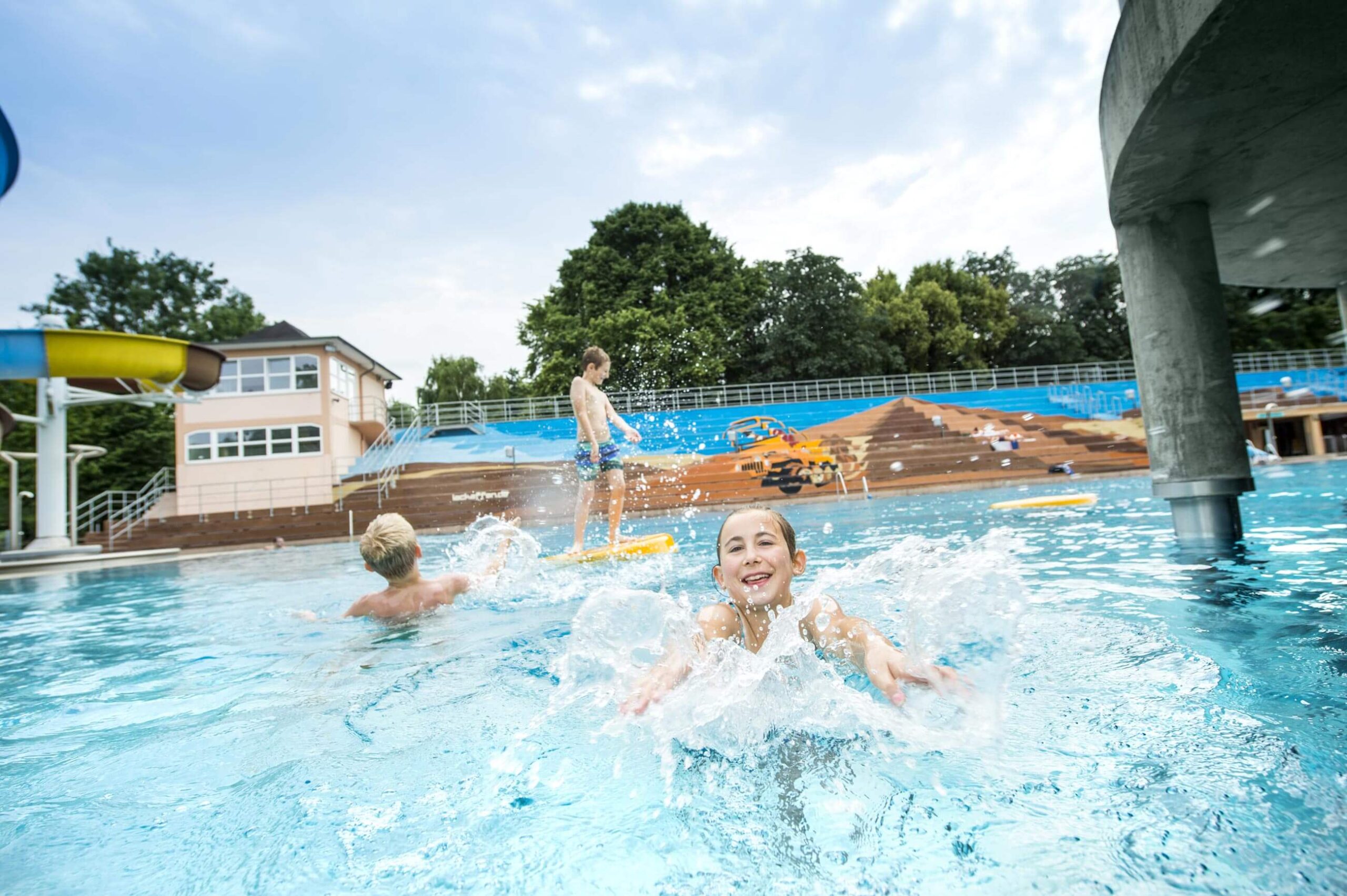 Ein lächelndes Mädchen spritzt im Freibadbecken mit Wasser, während im Hintergrund andere Kinder im Pool spielen.