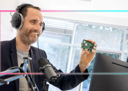 Nicolas Meseth mit Kopfhörern im Profil am Schreibtisch; er lächelt und hält eine kleine grüne Platine (Raspberry Pi) vor ein Mikrofon.