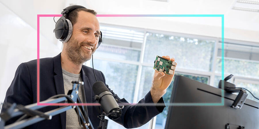 Nicolas Meseth mit Kopfhörern im Profil am Schreibtisch; er lächelt und hält eine kleine grüne Platine (Raspberry Pi) vor ein Mikrofon.