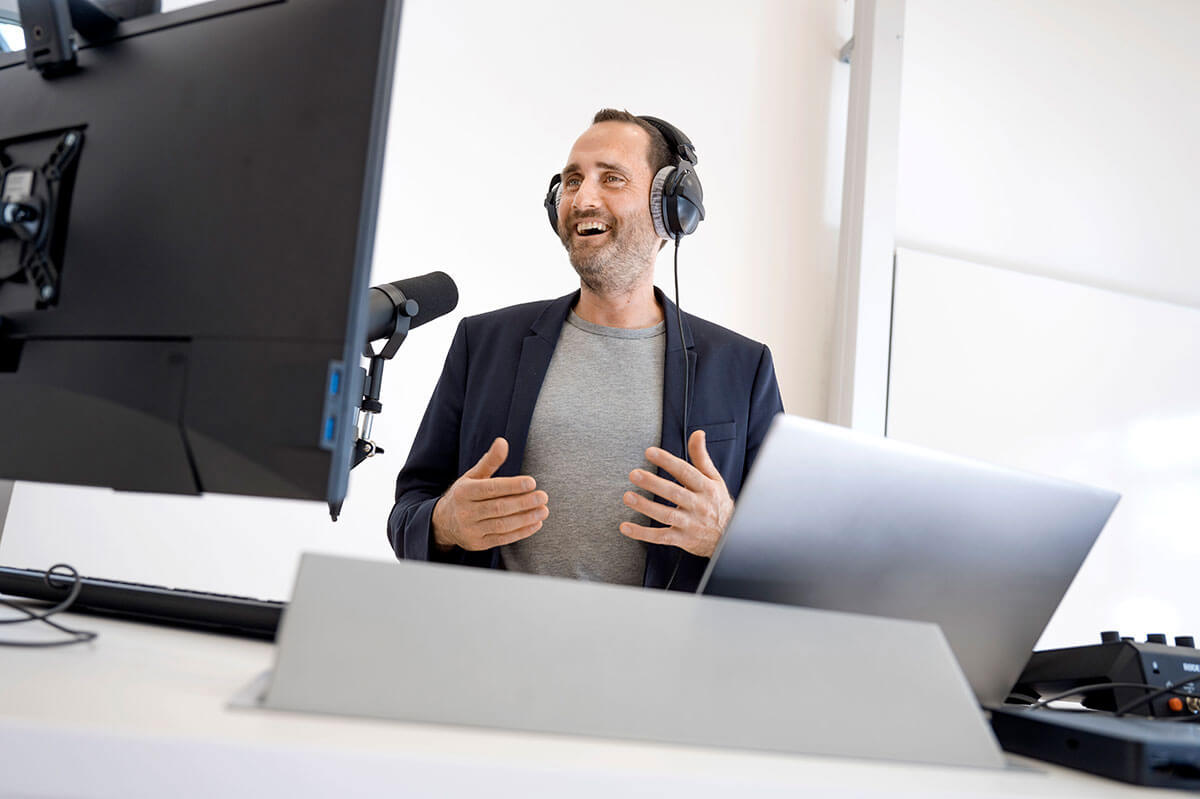 Nicolas Meseth spricht gestikulierend und lächelnd in ein Studiomikrofon; er trägt Kopfhörer, vor ihm steht ein Laptop.