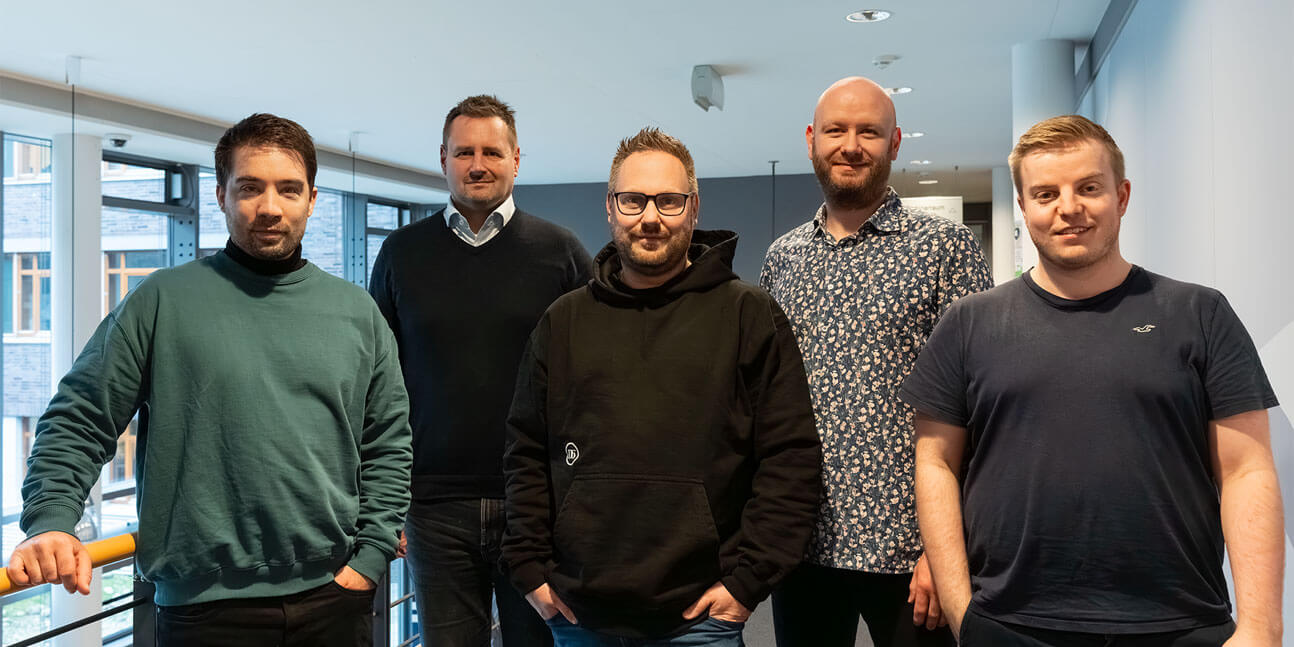 Fünf Männer stehen nebeneinander in einem hellen, modernen Büroflur und blicken in die Kamera.