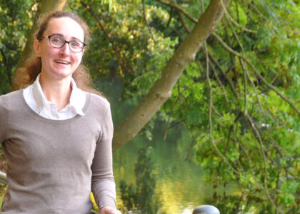 Porträt von Nina Kallmeier mit Brille und braunem Pullover, die lächelnd an einem Flussufer vor grünem Gebüsch steht.