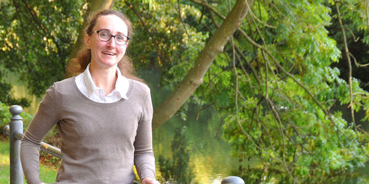 Porträt von Nina Kallmeier mit Brille und braunem Pullover, die lächelnd an einem Flussufer vor grünem Gebüsch steht.