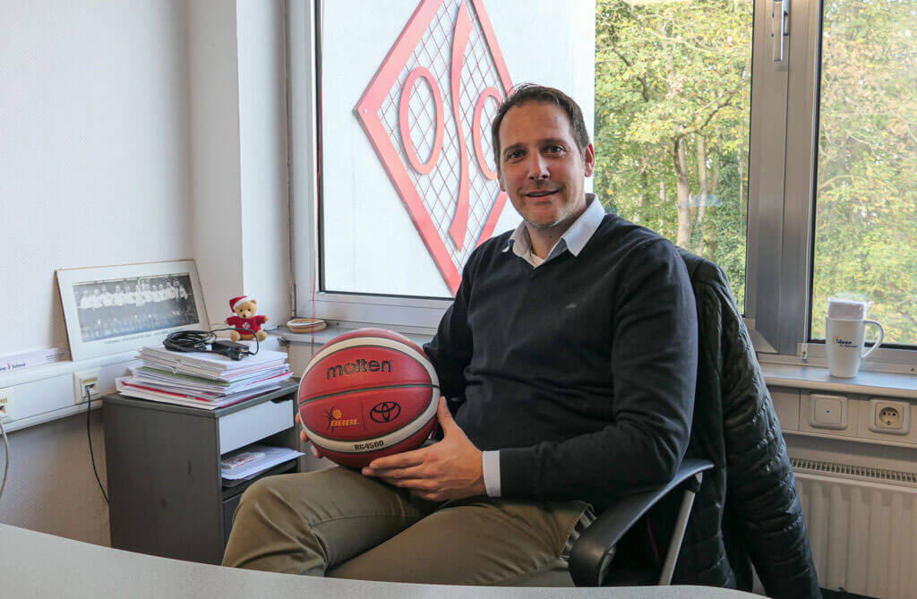 OSC-Geschäftsführer Sascha Bartsch sitzt lächelnd an seinem Schreibtisch im Büro und hält einen Basketball im Arm; im Hintergrund ist das rote OSC-Logo am Fenster zu sehen.