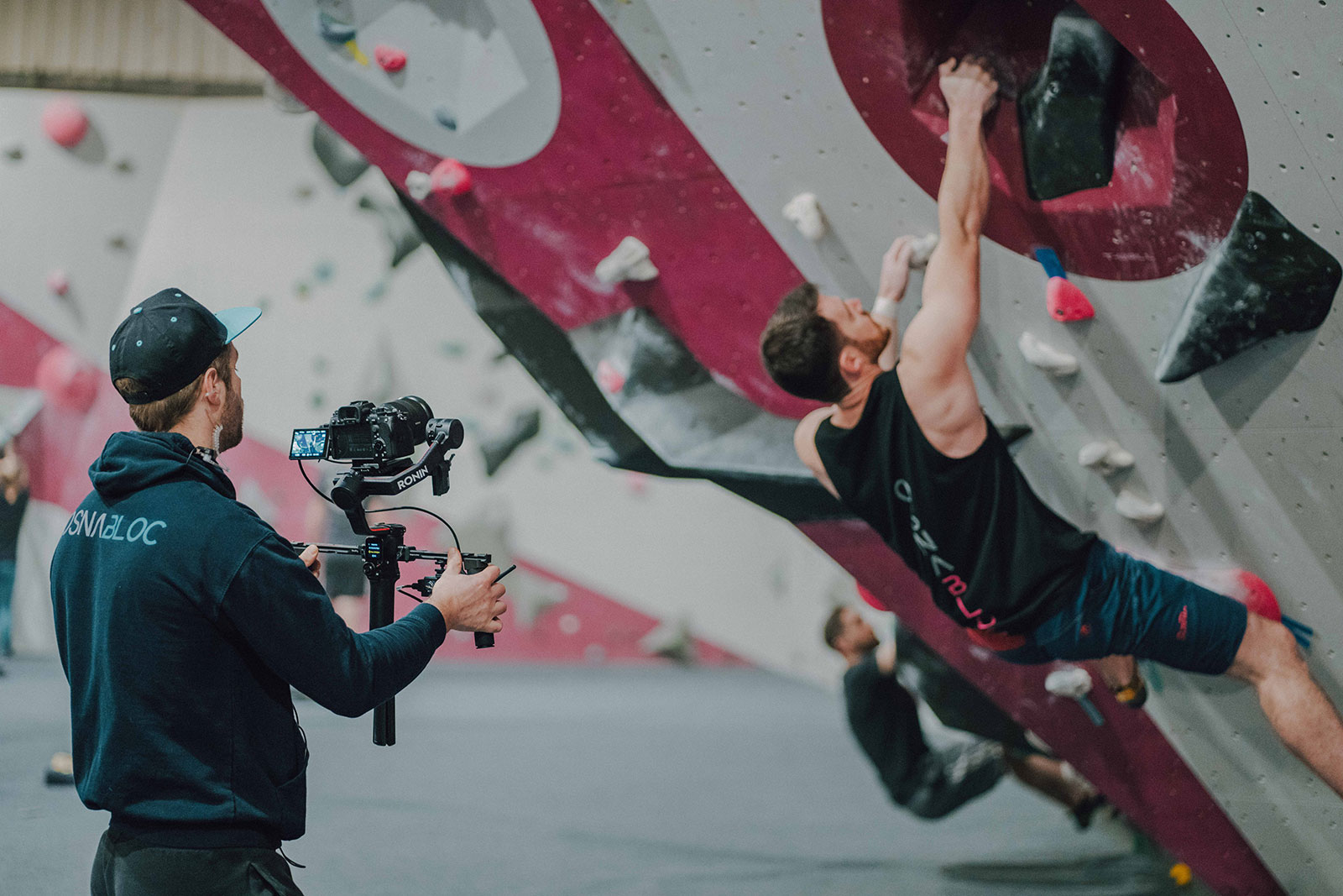 Ein Kameramann filmt einen Kletterer an einer steilen Wand in einer Boulderdach-Halle.