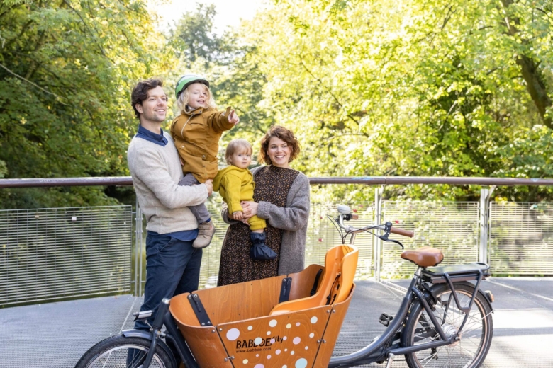 Eine lächelnde Familie mit zwei kleinen Kindern steht mit einem Lastenfahrrad auf einer Fußgängerbrücke.