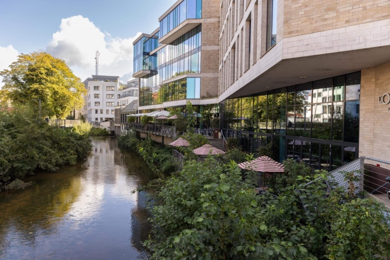 Moderne Gebäude mit Außengastronomie und Sonnenschirmen direkt an einem kleinen Flusslauf mit Bäumen.
