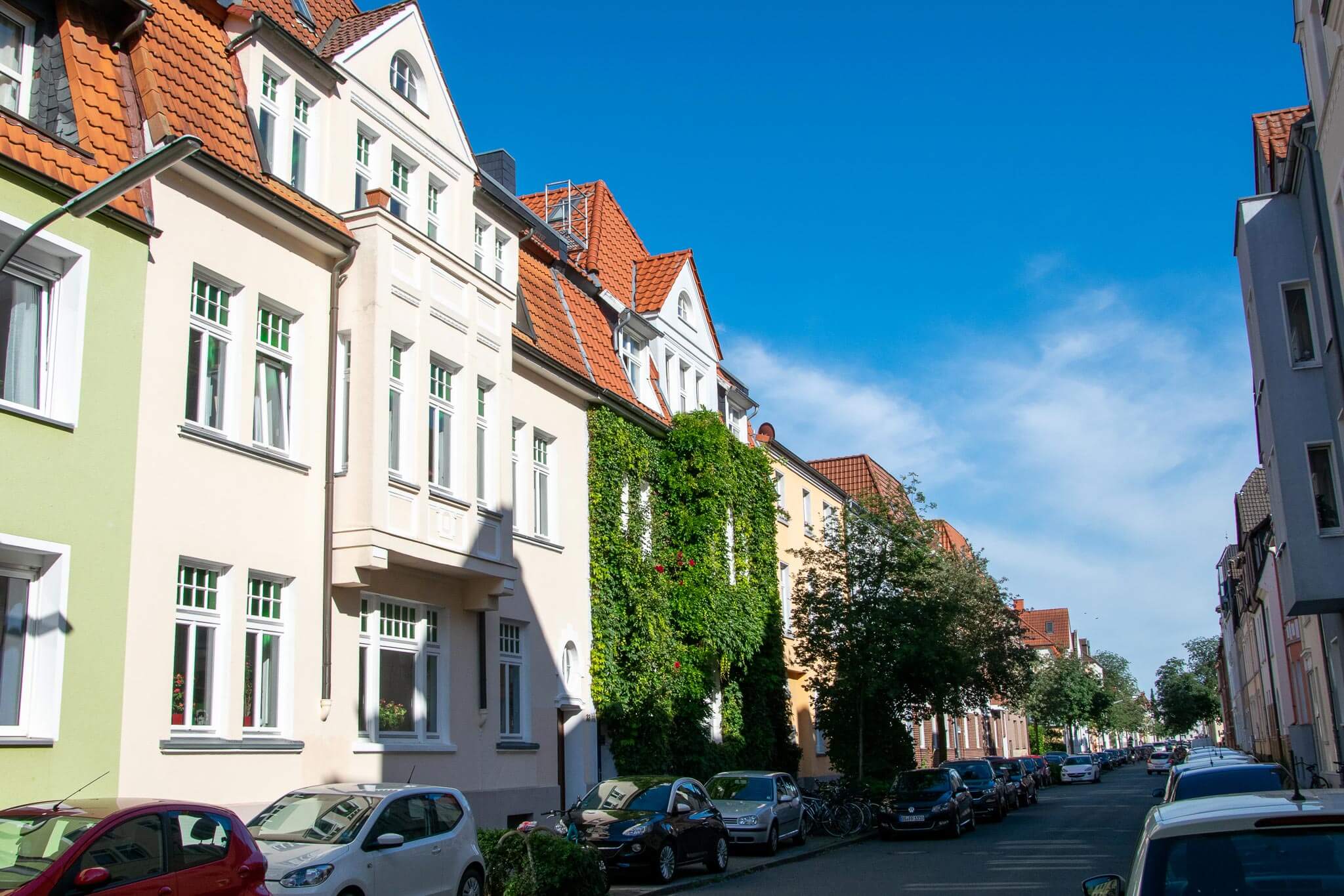 Sonnenbeschienene Wohnstraße mit mehrstöckigen Altbauten, einer efeuüberzogenen Fassade und geparkten Autos unter blauem Himmel.