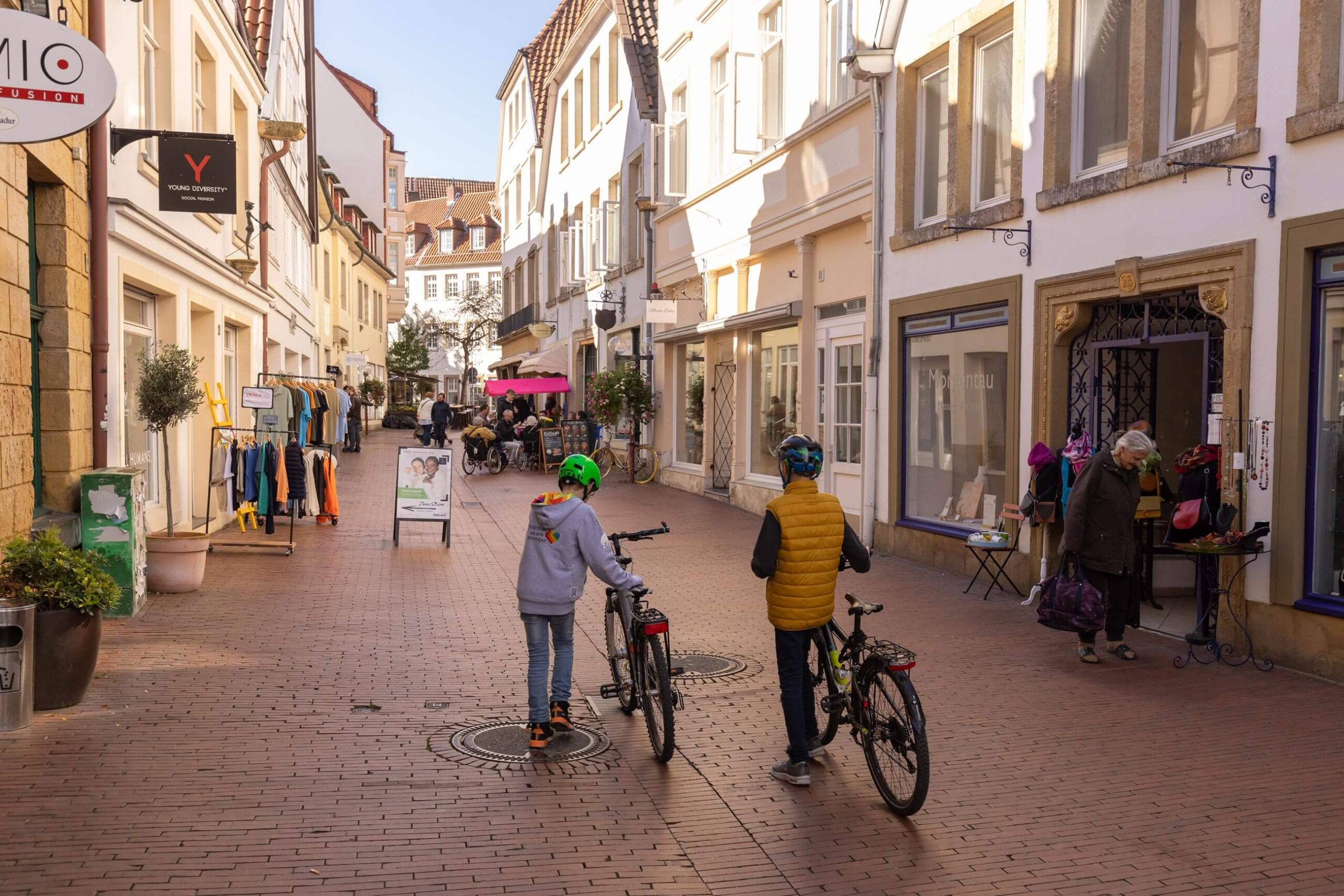 Zwei Kinder mit Helmen schieben ihre Fahrräder durch eine gepflasterte Fußgängerzone mit historischen Gebäuden und kleinen Geschäften.