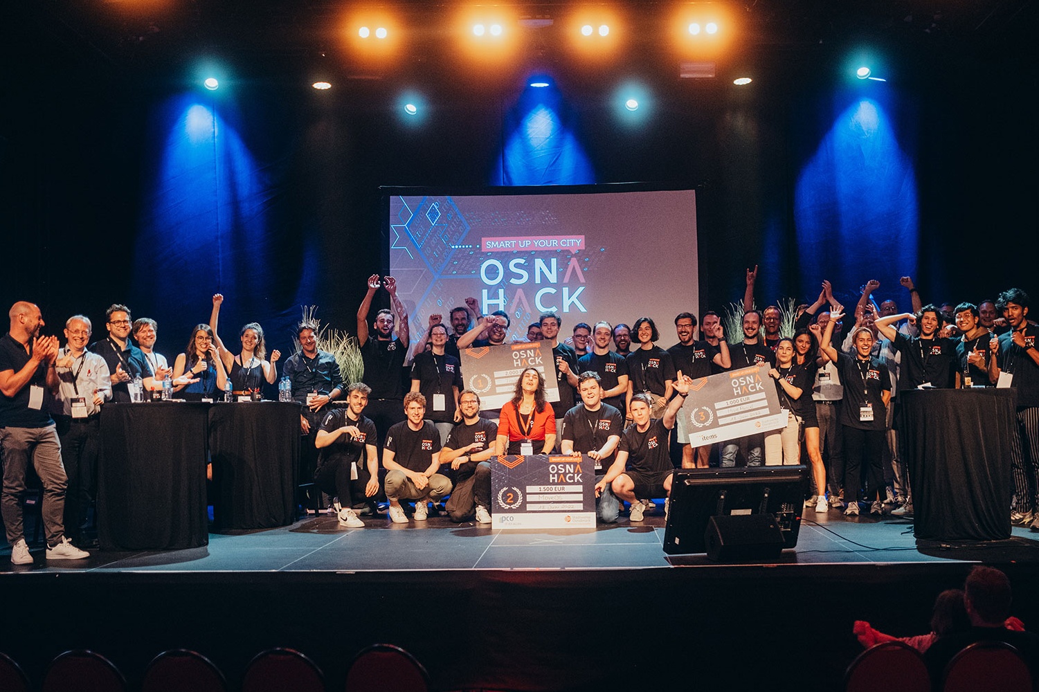 Viele Teilnehmer feiern mit großen Gewinnerschecks auf der beleuchteten Bühne der Hackathon-Veranstaltung OSNA HACK.