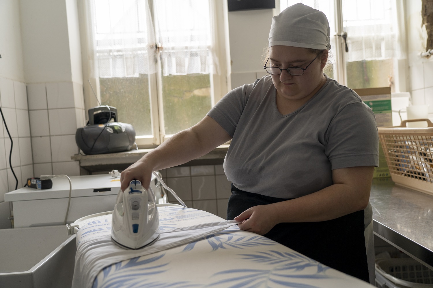 Eine Frau mit Kopftuch bügelt sorgfältig ein weißes Wäschestück auf einem Bügelbrett.