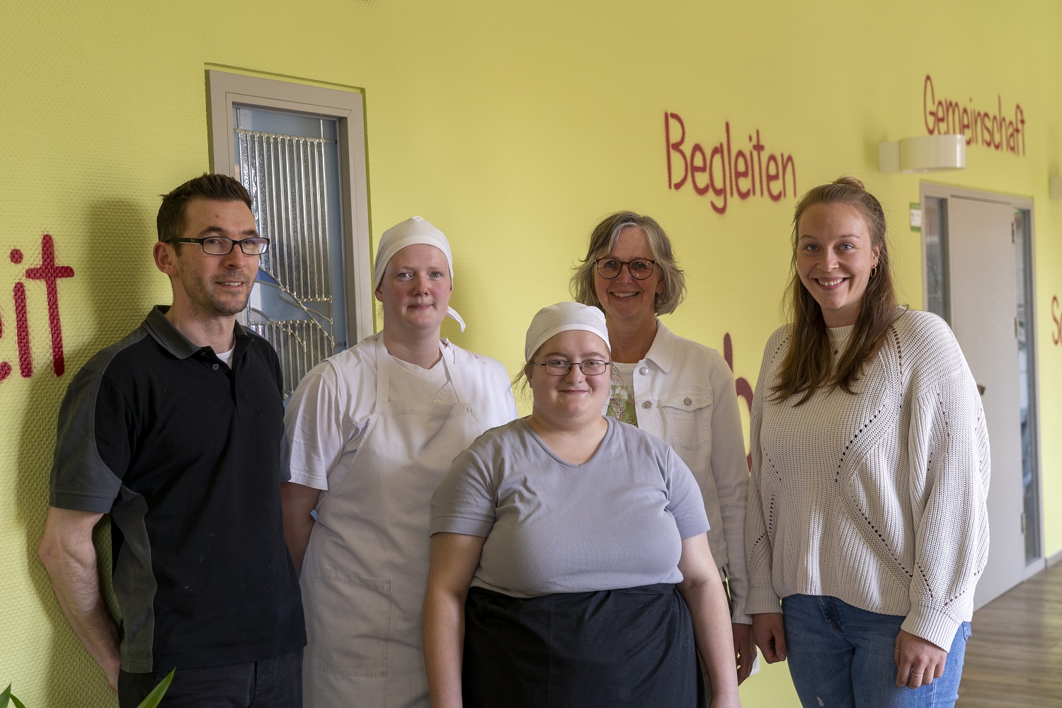 Fünf Personen stehen lächelnd nebeneinander vor einer gelben Wand mit der Aufschrift „Begleiten“.