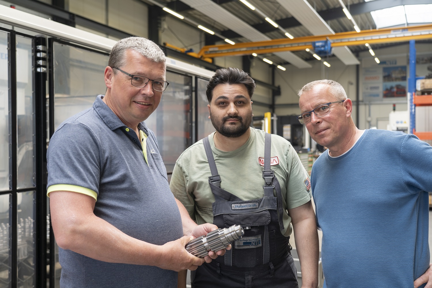 Drei Männer in einer Fabrikhalle stehen nebeneinander und betrachten gemeinsam ein metallenes Werkstück.