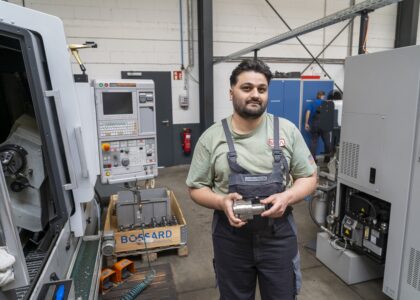 Ein Mann in Arbeitskleidung steht in einer Werkstatthalle und hält ein gefertigtes Metallteil in den Händen.