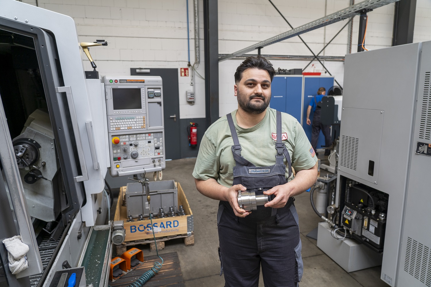 Ein Mann in Arbeitskleidung steht in einer Werkstatthalle und hält ein gefertigtes Metallteil in den Händen.