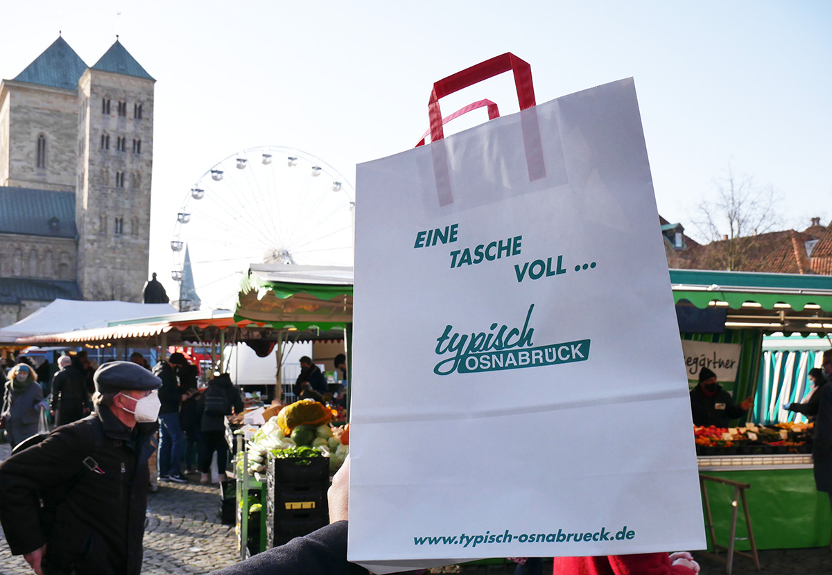 Eine weiße Papiertüte mit Aufdruck „typisch OSNABRÜCK“ wird auf einem geschäftigen Wochenmarkt gehalten.