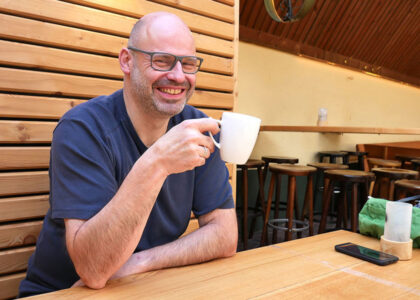 Pascal Rupp sitzt lächelnd mit Brille an einem Holztisch im Außenbereich und hält eine weiße Kaffeetasse in der Hand.