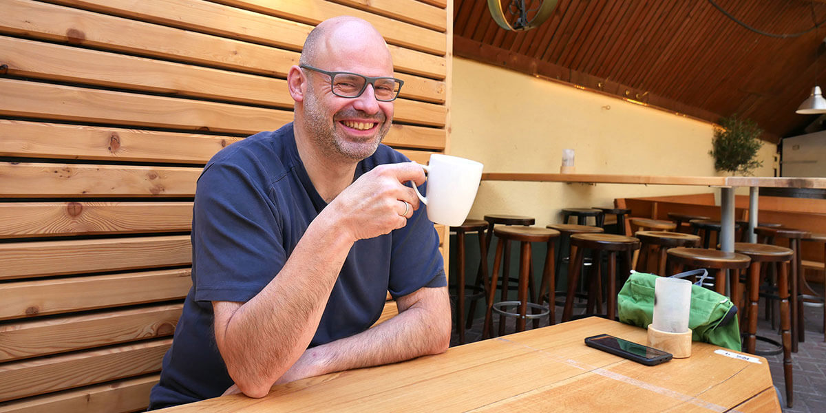 Pascal Rupp sitzt lächelnd mit Brille an einem Holztisch im Außenbereich und hält eine weiße Kaffeetasse in der Hand.