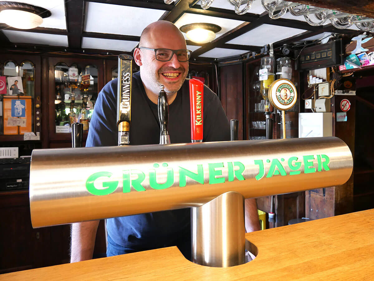 Pascal Rupp lächelt hinter der Zapfanlage einer Gastronomie, auf der in grüner Schrift ‚Grüner Jäger‘ steht.