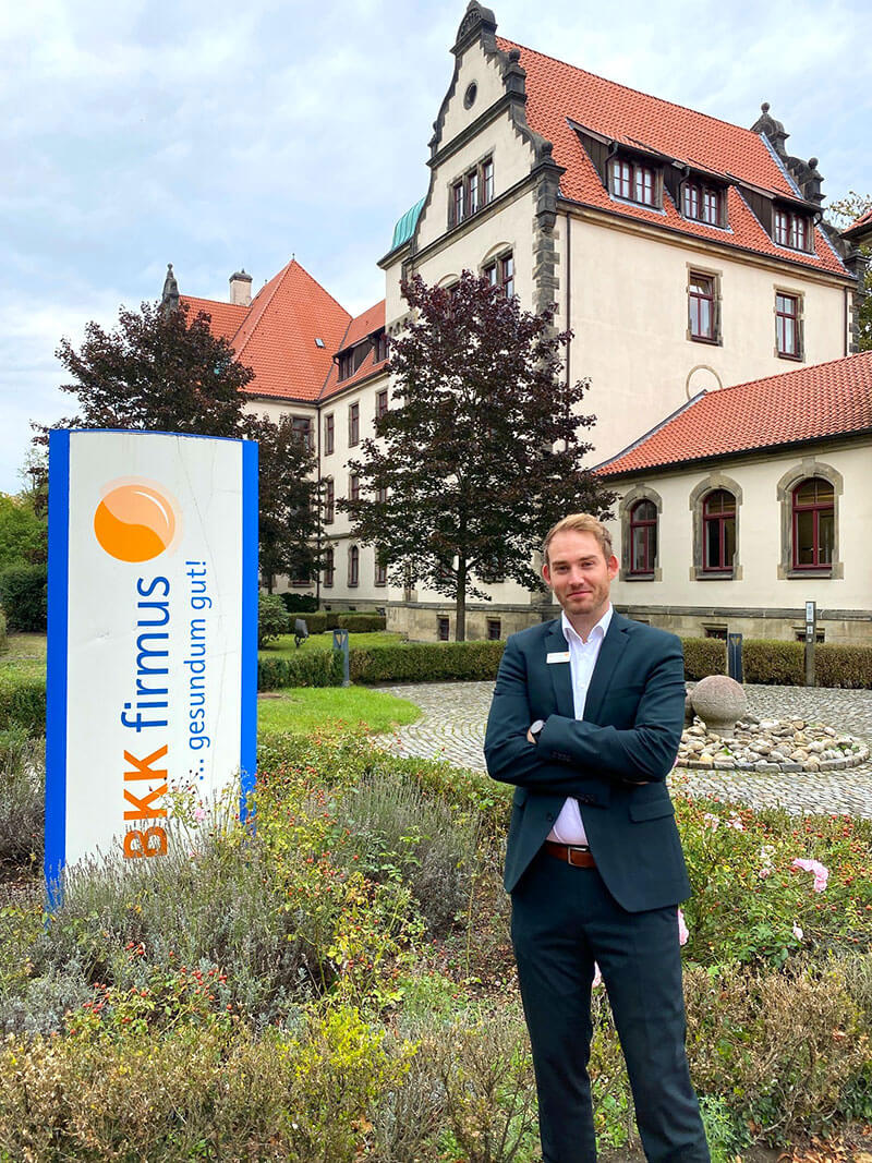 Patrick Schuecker im dunklen Anzug steht mit verschränkten Armen neben einer großen Werbesäule der BKK firmus vor einem historischen Gebäude.