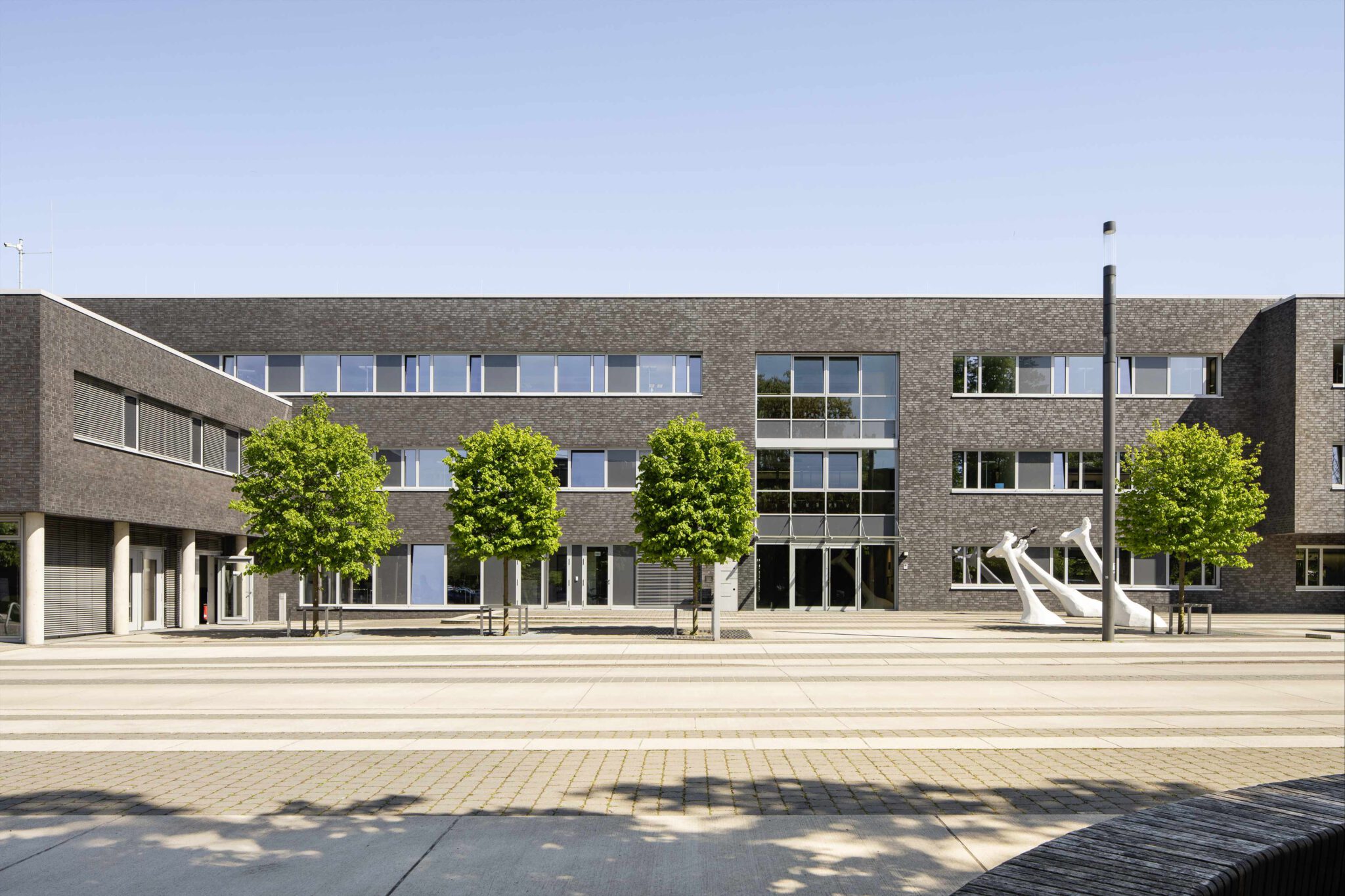 Moderne, dunkle Klinkerfassade eines Bürogebäudes mit großen Fensterfronten, Bäumen im Vorhof und Skulpturen.