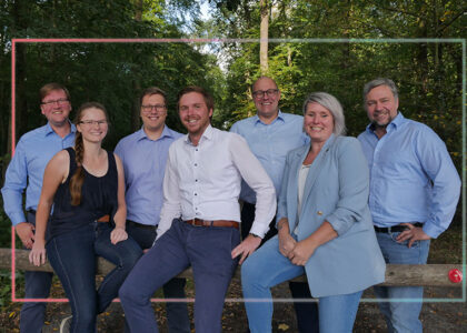 Gruppenfoto von sieben lächelnden Teammitgliedern vor einer bewaldeten Kulisse im Freien.