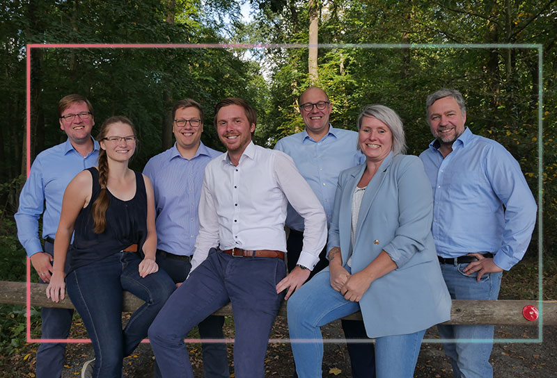 Gruppenfoto von sieben lächelnden Teammitgliedern vor einer bewaldeten Kulisse im Freien.
