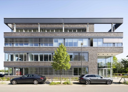 Mehrstöckiges Bürogebäude aus dunklem Klinker mit verglastem Eingangsbereich und geparkten Autos an der Straße.
