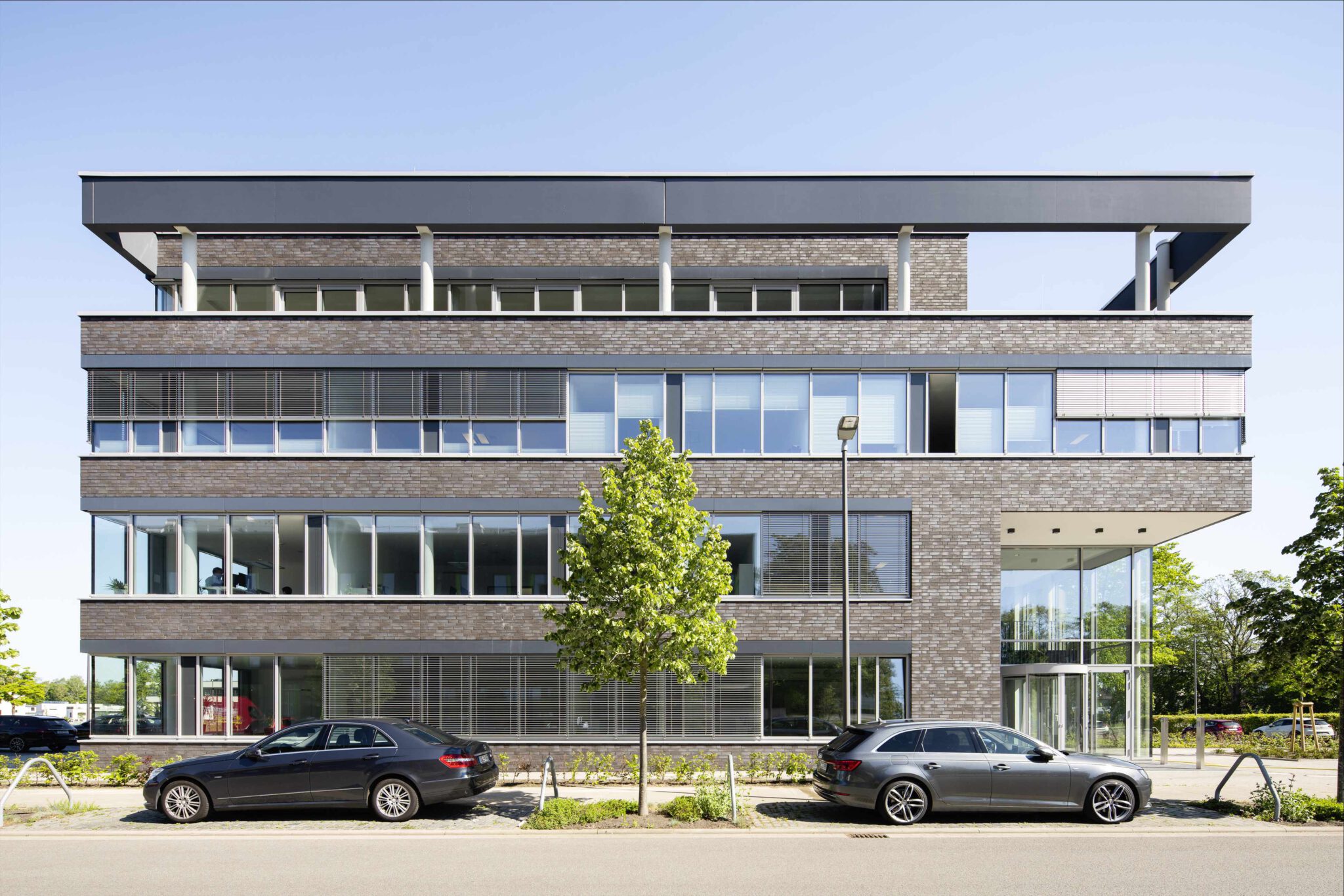 Mehrstöckiges Bürogebäude aus dunklem Klinker mit verglastem Eingangsbereich und geparkten Autos an der Straße.