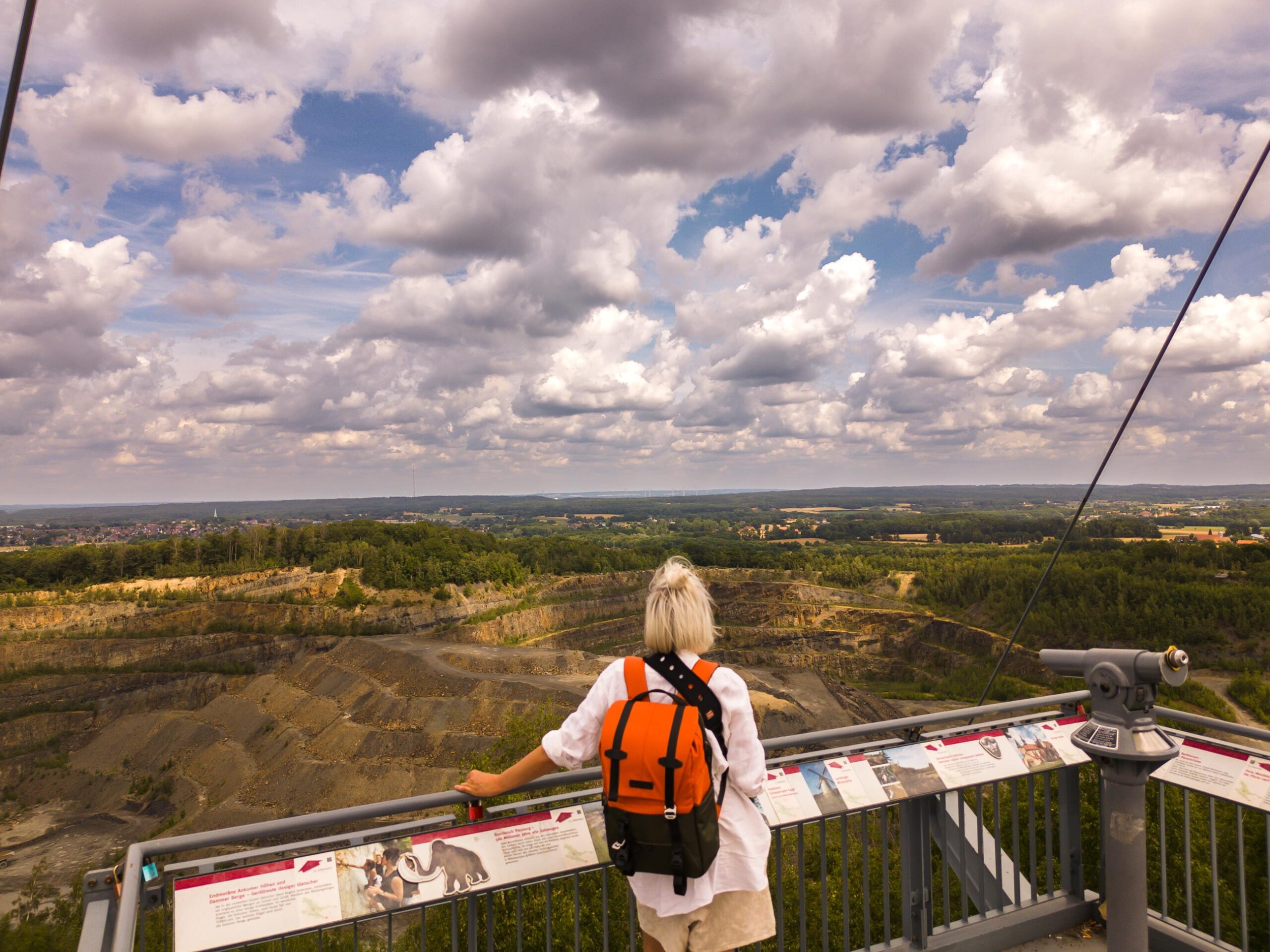 Blonde Frau mit orange-schwarzem Rucksack blickt von einer Aussichtsplattform mit Infotafeln auf einen riesigen Steinbruch unter bewölktem Himmel.