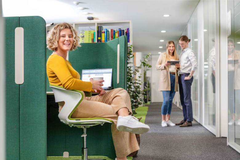 Eine Frau im gelben Oberteil sitzt lächelnd am Schreibtisch; im Hintergrund stehen zwei Personen im Flur.