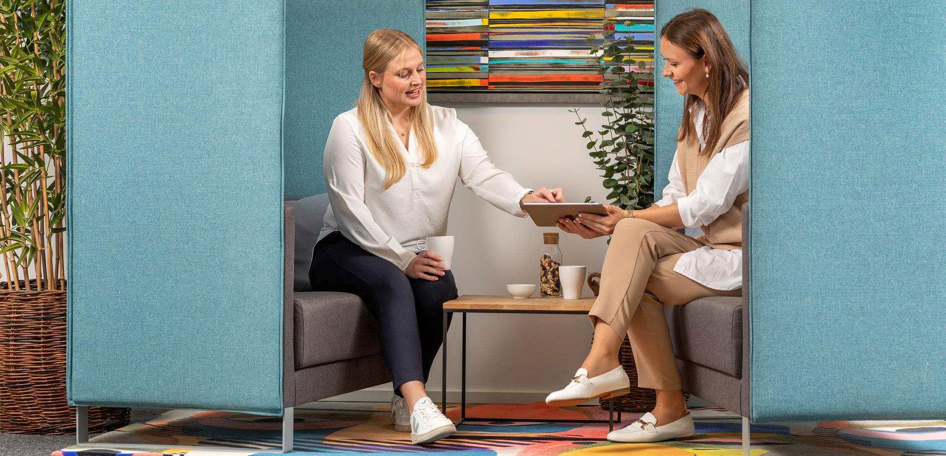 Zwei Frauen sitzen sich in einer blauen Besprechungskoje gegenüber und betrachten gemeinsam ein Tablet.