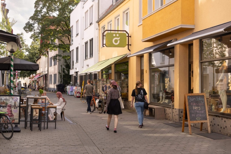 Menschen flanieren durch eine sonnige Einkaufsstraße mit gelben historischen Gebäuden, Straßencafés und gemütlichen Sitzgelegenheiten.