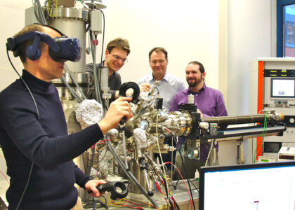 Wissenschaftler nutzt eine VR-Brille und Controller an einer komplexen Laboranlage, während drei Kollegen im Hintergrund zusehen.