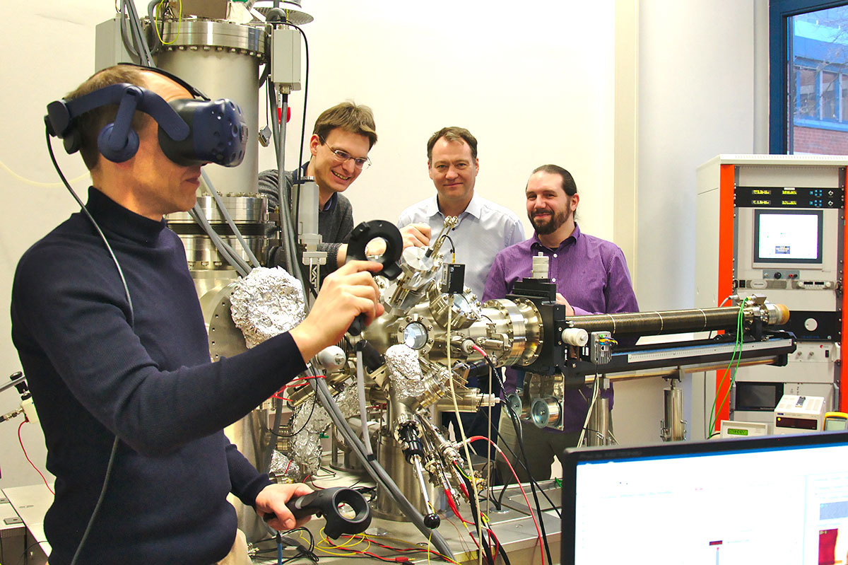 Wissenschaftler nutzt eine VR-Brille und Controller an einer komplexen Laboranlage, während drei Kollegen im Hintergrund zusehen.