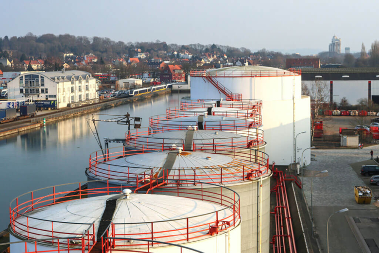 Große, runde Öltanks mit roten Geländern an einem Hafenkanal in der Vogelperspektive.