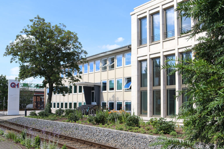 Gebäudekomplex mit Treppenaufgang, bepflanztem Vorgarten und Bahngleisen im Vordergrund.