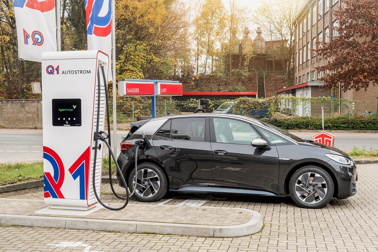 Dunkelgraues Elektroauto lädt an einer weißen Q1-Schnellladesäule auf einem Tankstellengelände.