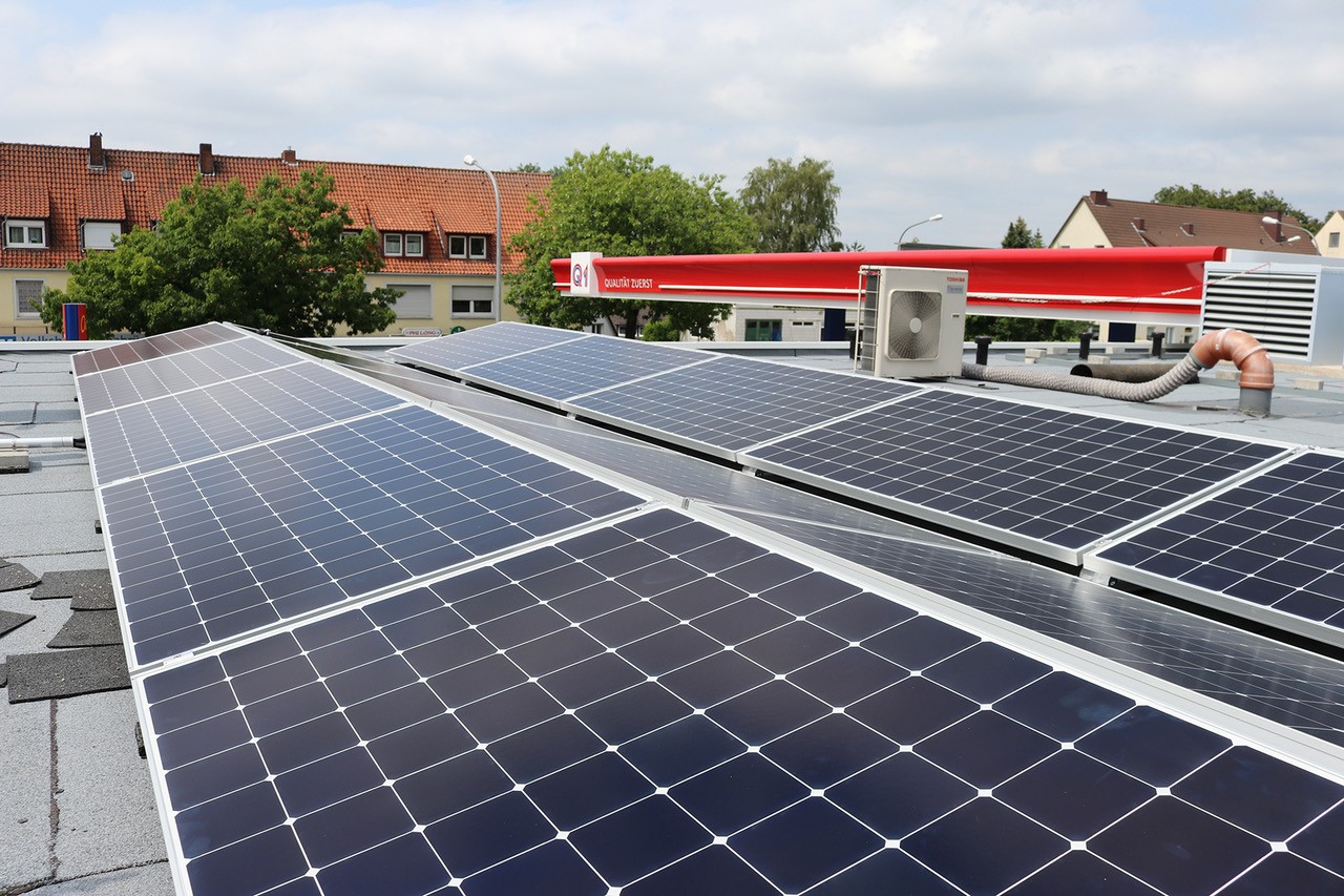 Aufreihte Solarmodule auf dem Flachdach eines Q1-Tankstellengebäudes mit Wohnhäusern und Bäumen im Hintergrund.