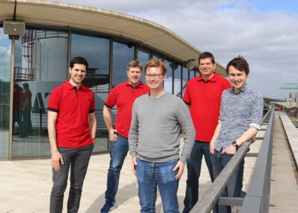 Fünf lächelnde Männer stehen auf der Dachterrasse eines modernen Gebäudes, drei davon tragen rote Poloshirts.