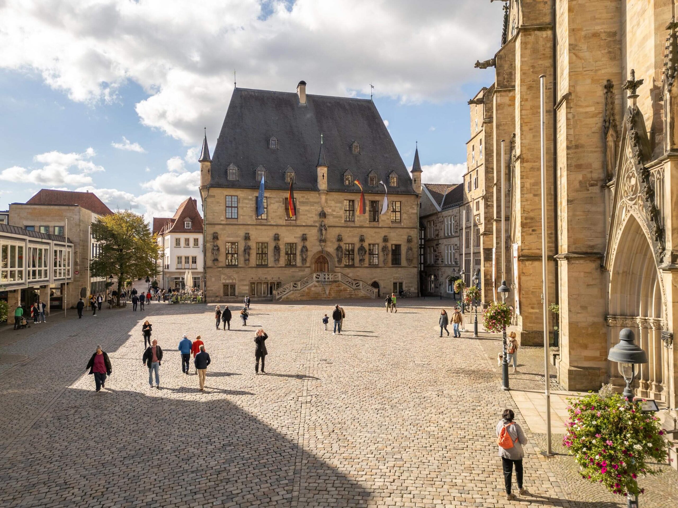 Historisches Rathaus Osnabrück mit Freitreppe an einem gepflasterten Marktplatz mit Passanten unter leicht bewölktem Himmel.