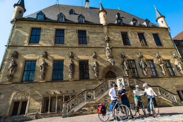 Vier Personen mit Fahrradhelmen betrachten eine Straßenkarte vor der Freitreppe eines historischen Rathausgebäudes.