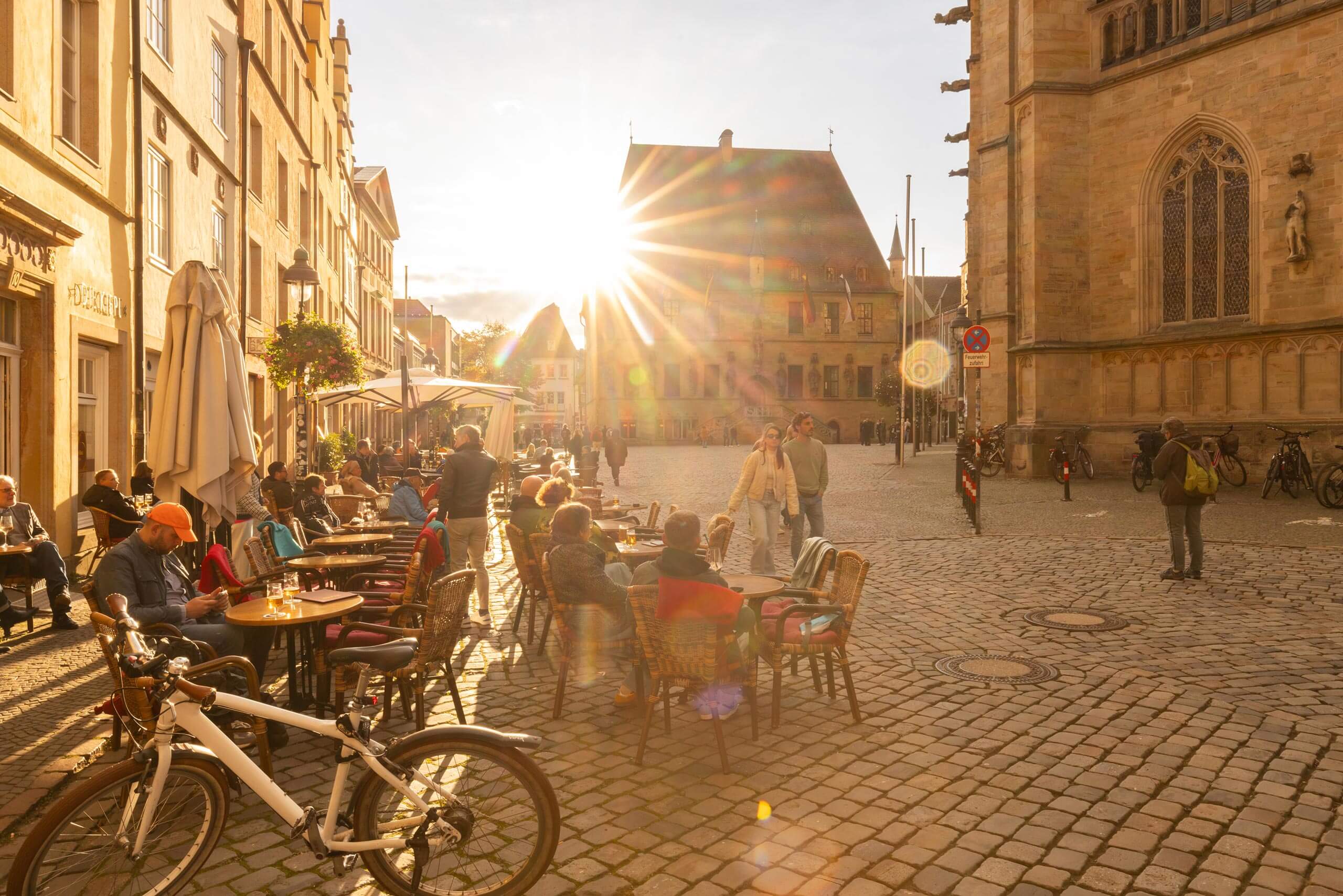 Eine historische Altstadt mit Cafés und Tischen im warmen Abendlicht der tiefstehenden Sonne, die strahlende Lichtstrahlen wirft.