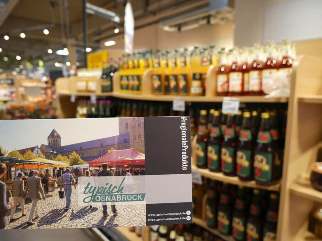 Eine Hand hält eine Postkarte mit der Aufschrift „typisch OSNABRÜCK“ vor ein Supermarktregal mit Getränken.