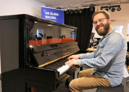 Ein Mann mit Bart und Brille sitzt lächelnd an einem schwarzen Klavier und spielt darauf.
