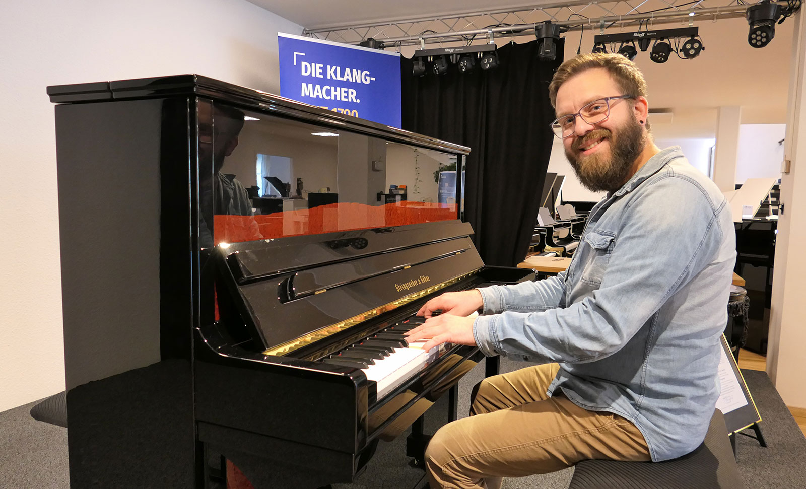 Ein Mann mit Bart und Brille sitzt lächelnd an einem schwarzen Klavier und spielt darauf.