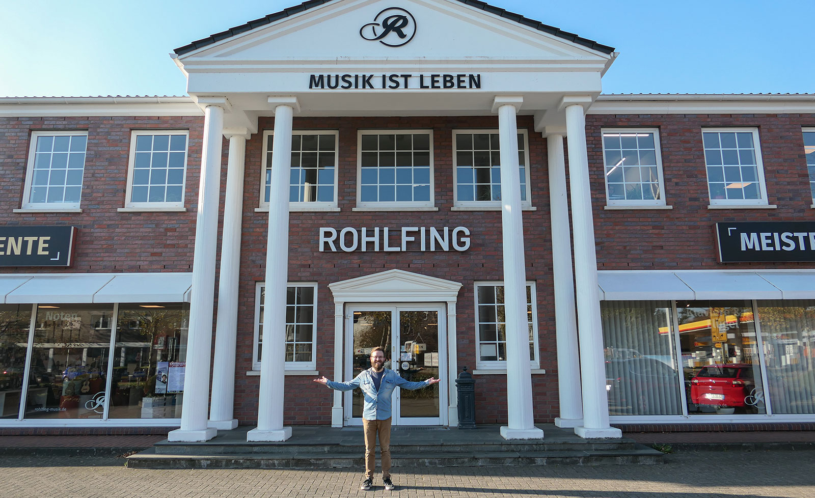 Ein Mann steht mit ausgebreiteten Armen vor einem Gebäude mit Säulen und der Aufschrift „Rohlfing“.