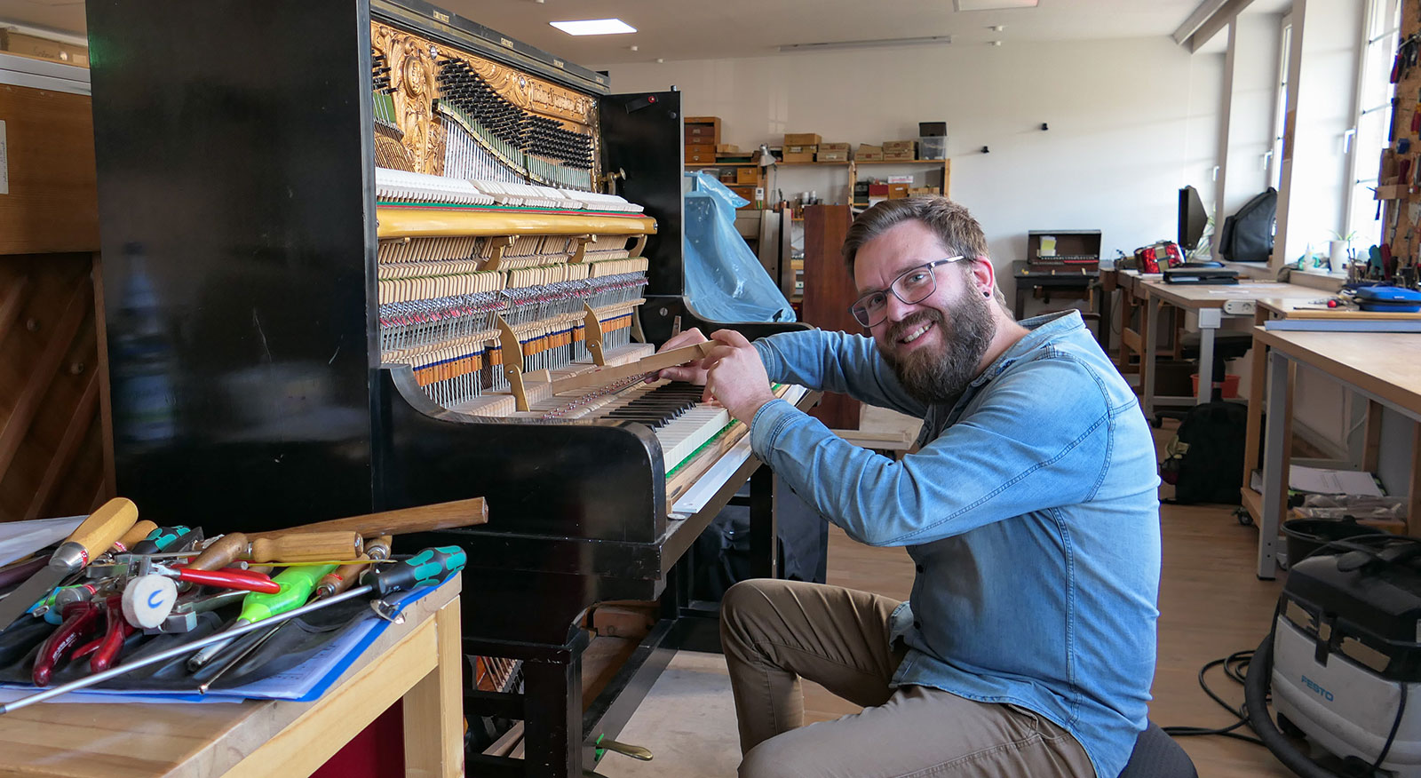 Ein Mann arbeitet in einer Werkstatt lächelnd an der Mechanik eines geöffneten Klaviers.