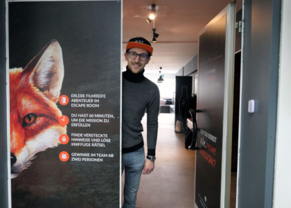 Mann schaut hinter einer geöffneten Tür hervor, die mit einem Fuchs-Motiv und Hinweisen zu einem Escape-Room bedruckt ist.